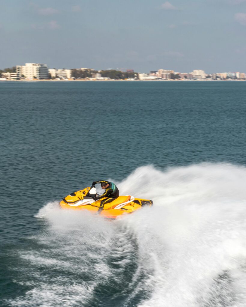 An adult wearing a helmet rides a jet ski, creating a large splash on the clear ocean near a coastal city.