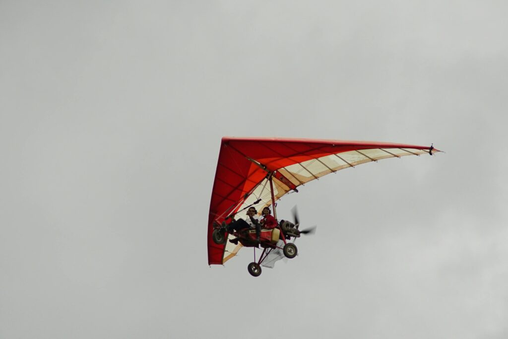 A vibrant red powered hang glider with two pilots soaring in an overcast sky.